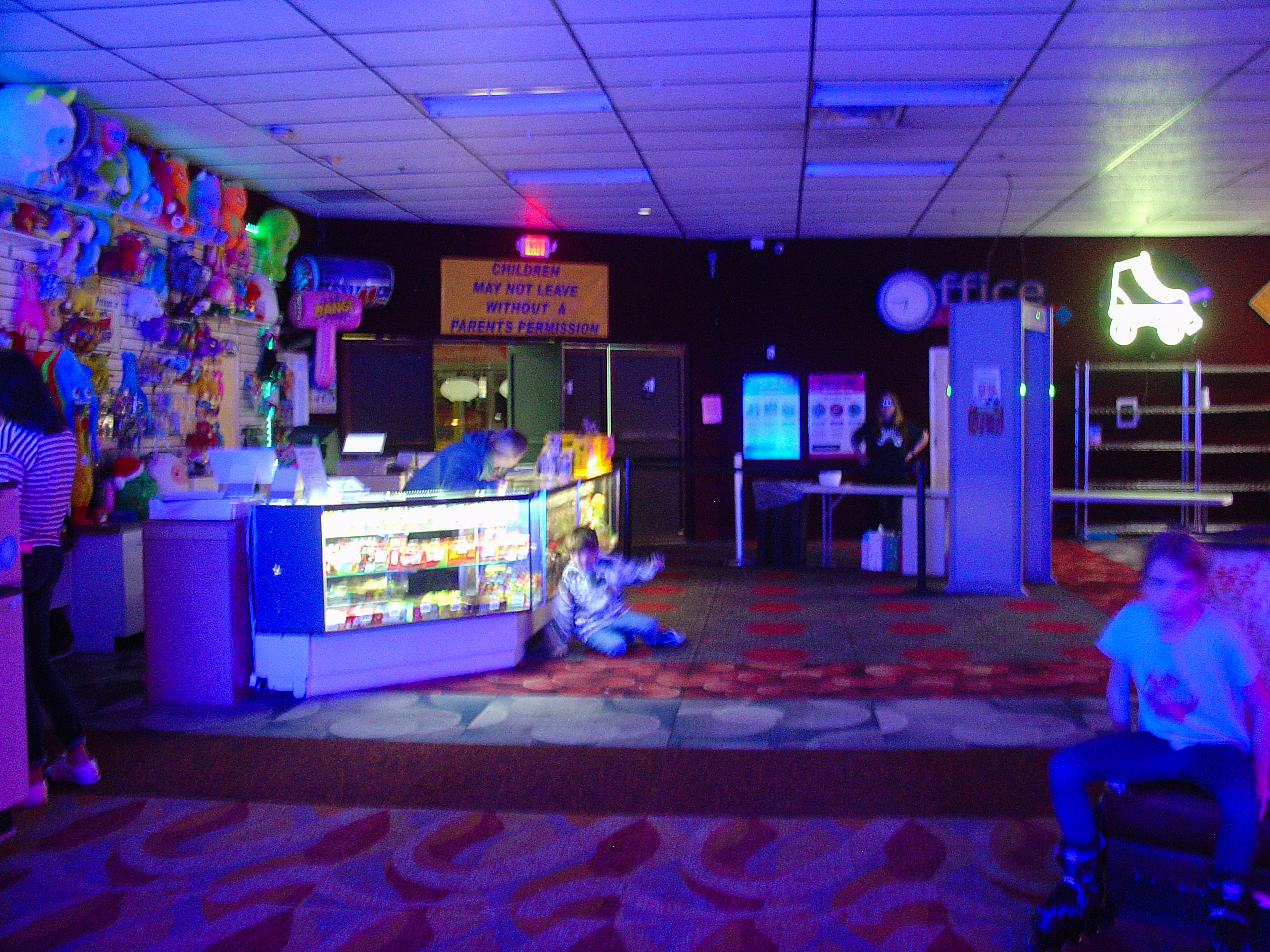 A roller rink at night lit in pink and blue neon, a snack counter and a glowing roller-skate sign, kids scattered across the patterned carpet