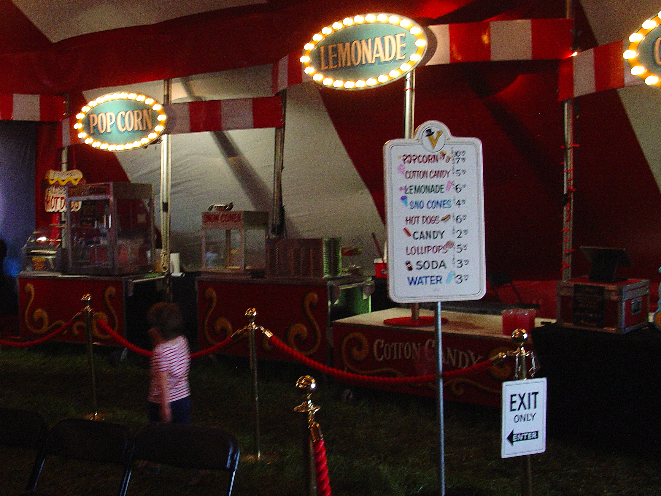 The inside of a striped carnival tent, lit popcorn and lemonade signs over a concession stand, a small child by a red rope