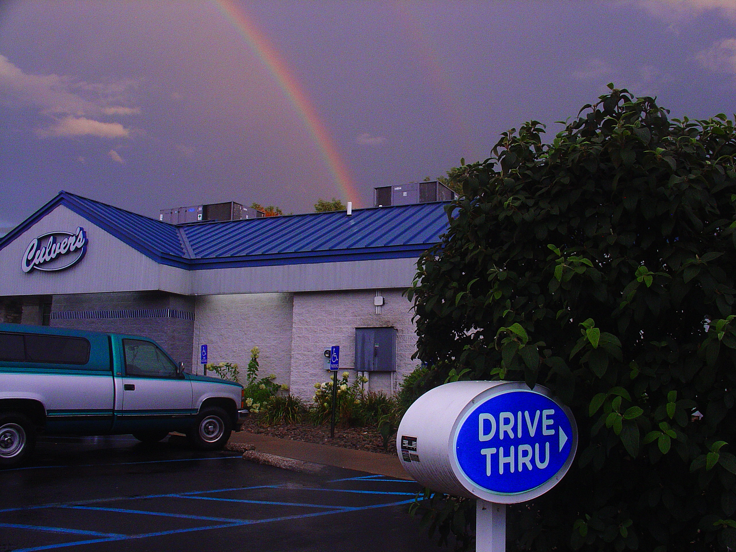 A double rainbow over a Culver's restaurant with a blue roof, a teal pickup in the lot, color fringing visible along the truck's edges