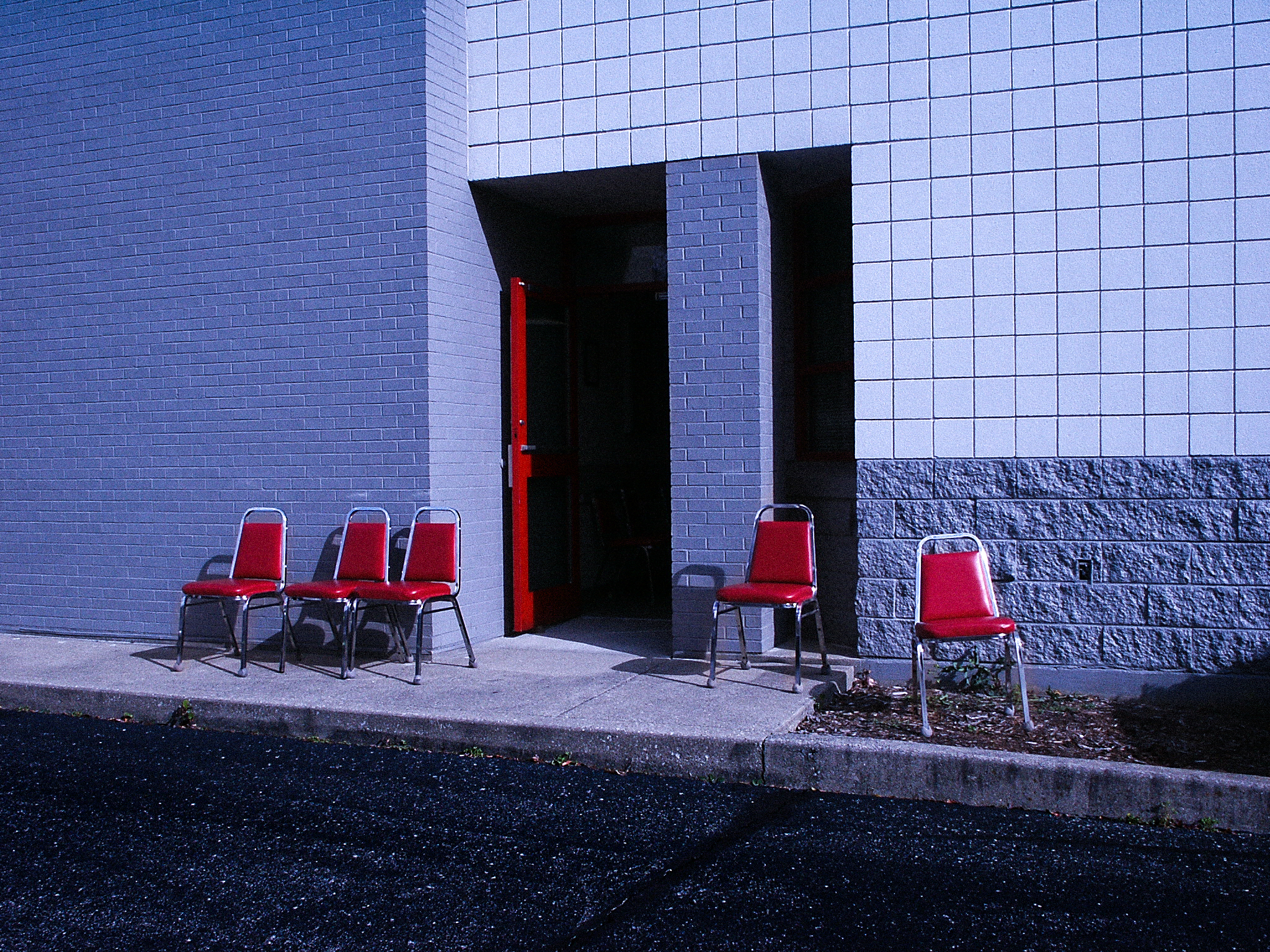 Red stacking chairs set out on the pavement beside a propped red door in a two-tone brick wall, hard daylight