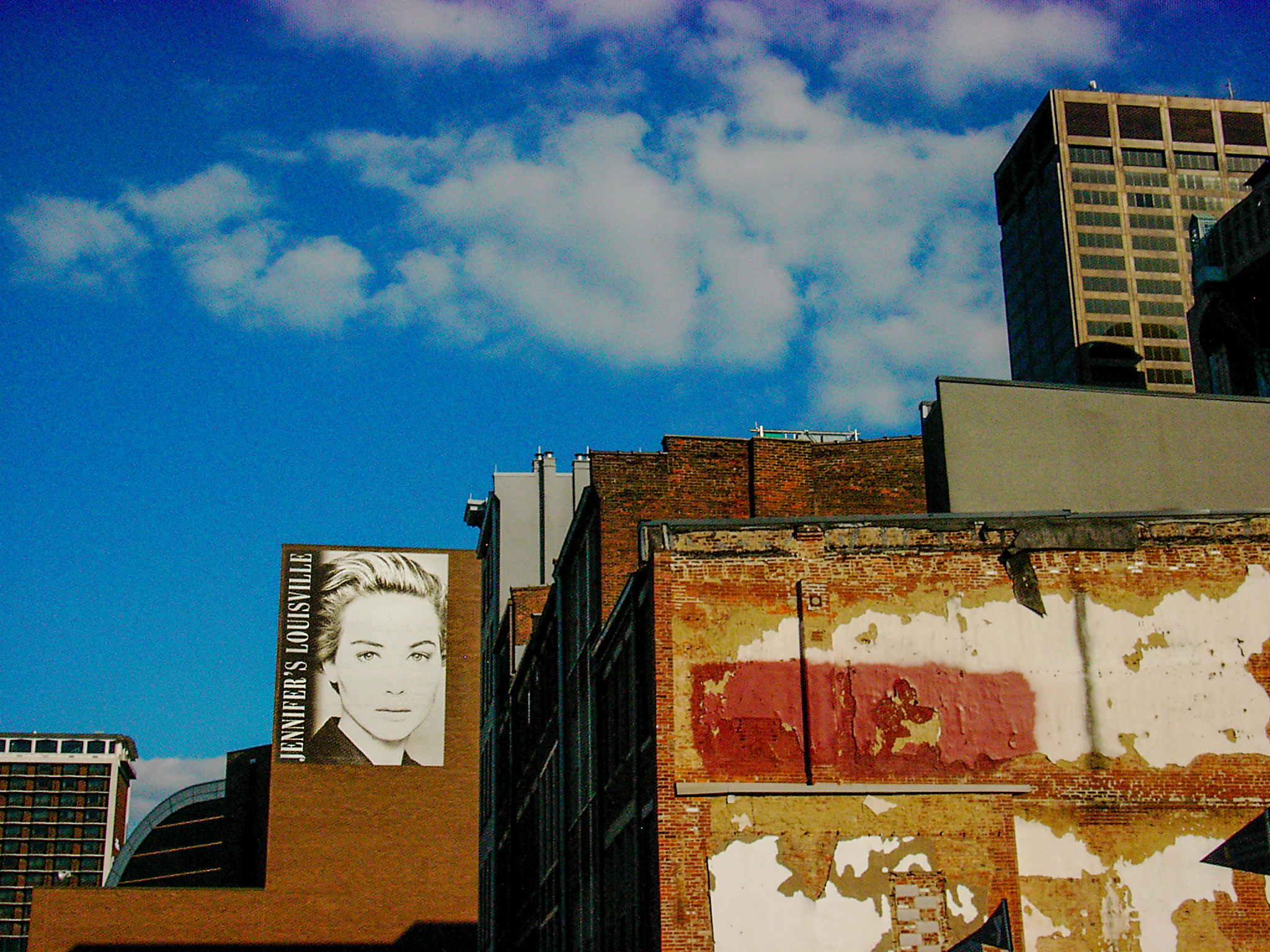 A sunlit city wall with peeling red paint and a pasted face, blue sky and a tall building behind it
